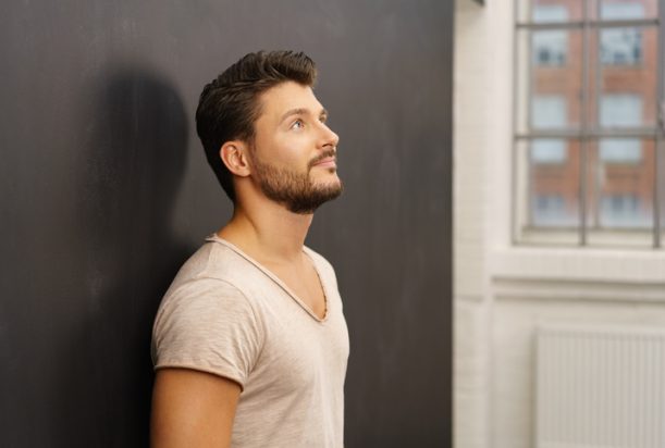 Thoughtful man standing leaning against a dark grey wall looking up with a contemplative expression